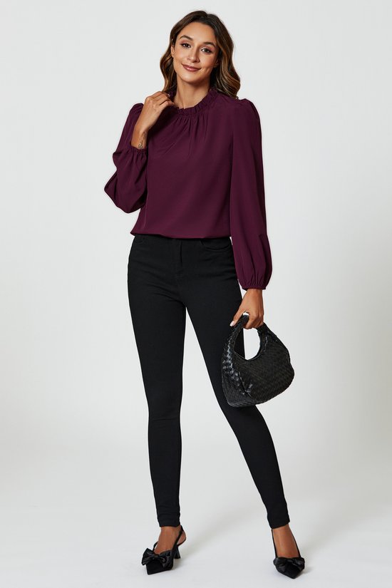Vrouw in pruimkleurige blouse met ruches en hoge hals, ballonmouwen en zwarte skinny jeans, met zwarte tas.
