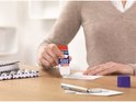 Une personne utilisant un bâton de colle Tesa Easy Stick avec capuchon bleu et base rouge pour coller du papier blanc.