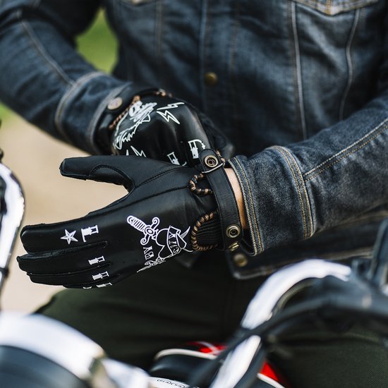 Zwarte motorhandschoenen met witte tatoeage-achtige prints van een kruisend zwaard en hart, een bliksemschicht, sterren en tekst. Mannenhanden trekken de handschoenen aan.