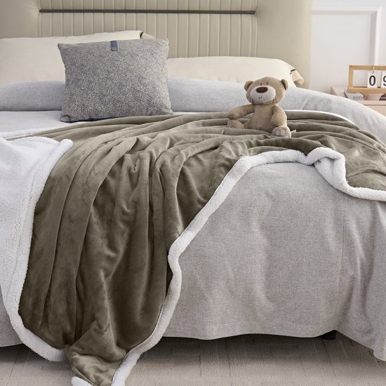 Taupe fleece plaid met witte sherpavoring, gedrapeerd over een bed met een teddybeer.