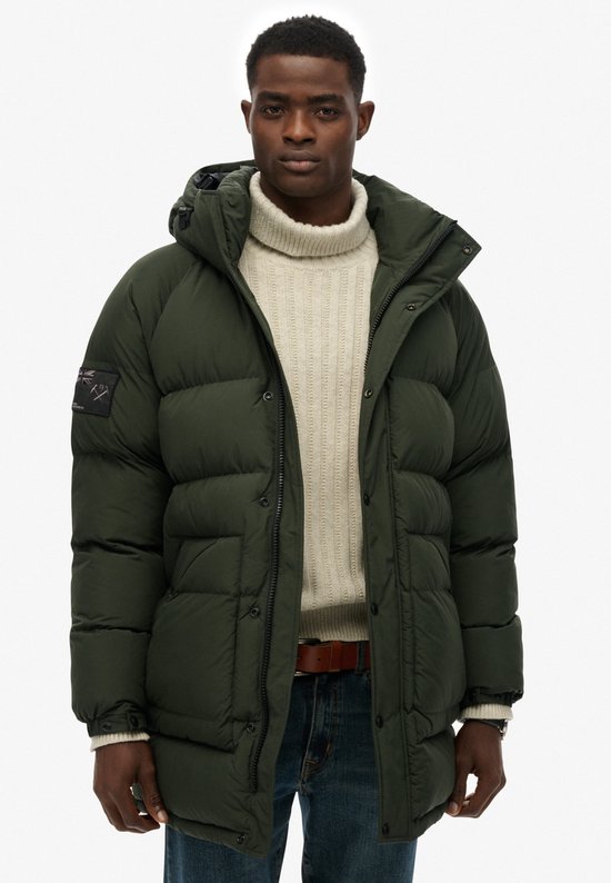 Een man draagt een lange, donkergroene, gewatteerde parka met capuchon, ritssluiting en knopen.
