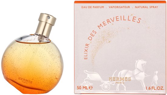 Ronde, oranje Elixir des Merveilles eau de parfum flacon met gouden dop, bezaaid met gouden glitters. Naast de flacon een grijze doos met oranje rand.
