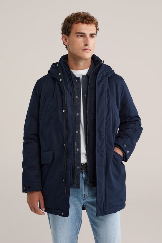 Man met donkerblauwe parka, lichte jeans en witte sneakers. Parka met capuchon, ritssluiting en klepzakken.
