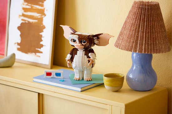 Een LEGO Ideas Gizmo figuur op een blauw boek, naast een gele mok en een 3D-bril. De figuur is bruin en wit met grote oren.