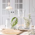 Une table dressée avec un verre à pied portant une bougie, un verre avec une feuille verte, un verre à pied et un verre avec des fleurs.