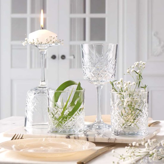 Une table dressée avec un verre à pied portant une bougie, un verre avec une feuille verte, un verre à pied et un verre avec des fleurs.