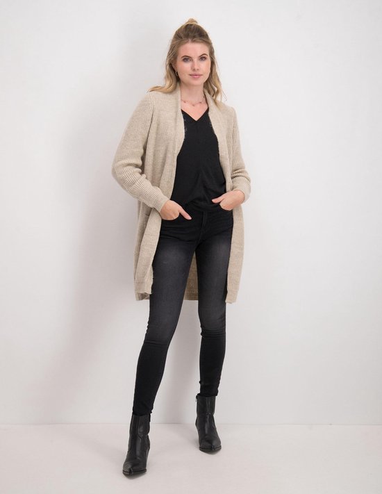 Model in een lange beige vest en zwarte skinny jeans.