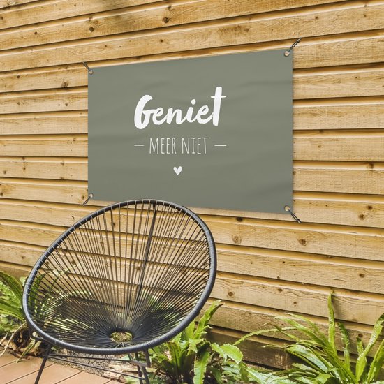 Donkergroene banner met witte tekst 'Geniet - Meer Niet -' en een hartje, hangend aan een houten schutting naast een zwarte stoel.
