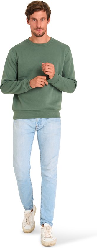 Man in mosgroene sweater en lichtblauwe jeans. Hij stapt naar voren met zijn handen bij zijn polsen.