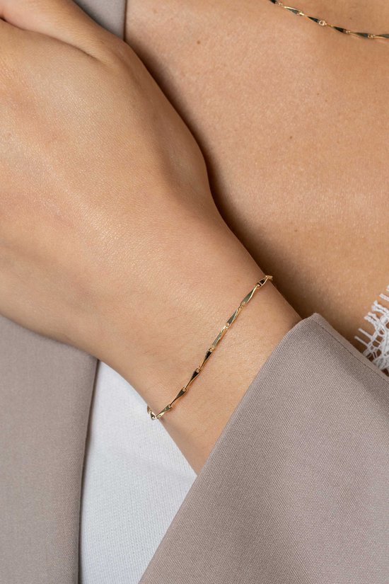 Close-up van een model met een gouden schakelarmband met platte, pijl-vormige schakels om de pols. Ze draagt een bruin colbert.