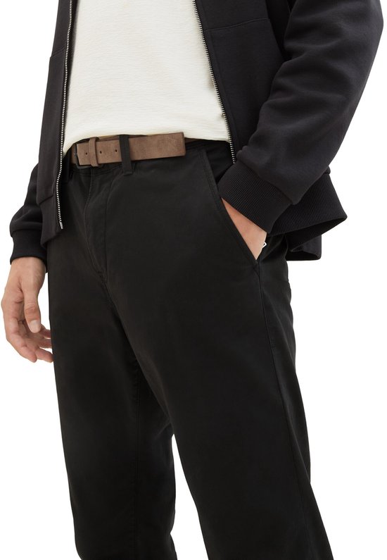 Man in close-up, draagt een zwarte chino en een bruine riem, met een hand in de zak.