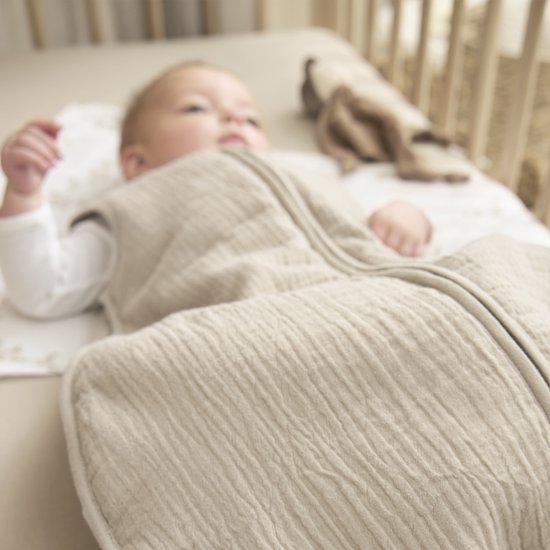Zicht op een baby in een zandkleurige, gevoerde slaapzak. De slaapzak heeft een gebreide structuur.