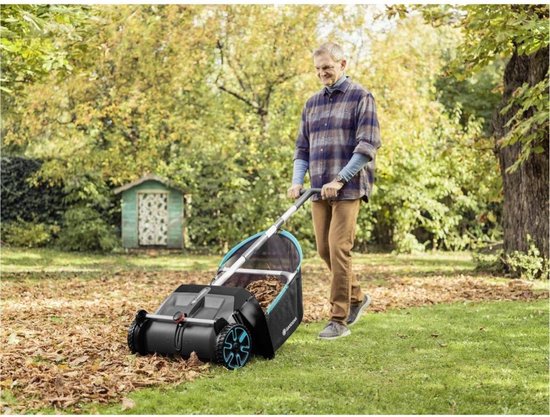 Man duwt Gardena bladverzamelaar over een grasveld bezaaid met bladeren, met een klein huisje op de achtergrond.