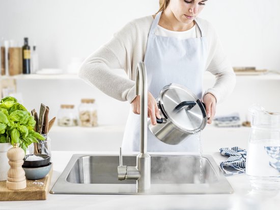 Vrouw met schort bij de gootsteen die een zilverkleurige kookpot afspoelt onder de kraan.