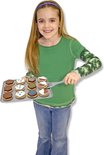 Een meisje met lang blond haar houdt een bakplaat met houten koekjes vast. Ze draagt een groene trui en spijkerbroek.