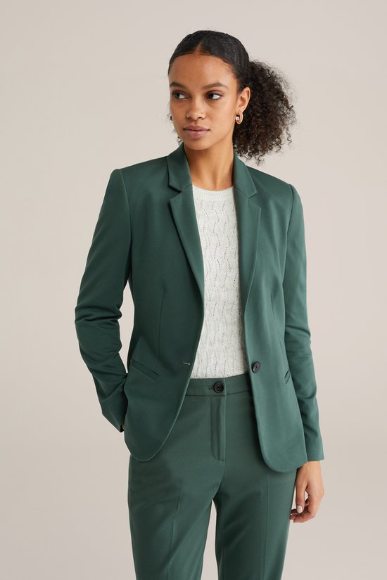 Model met de handen in de zakken van een donkergroene blazer en broek.