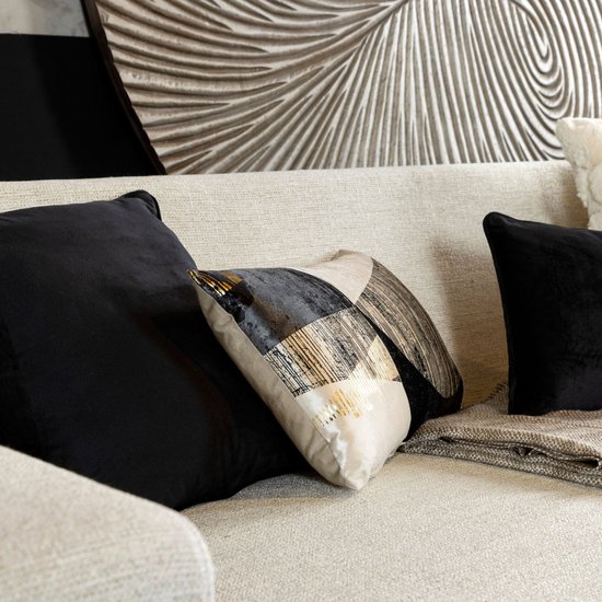 Een decoratief kussen met geometrische patronen in zwart, goud en beige, naast twee effen zwarte fluwelen kussens op een lichte bank.