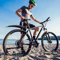 Man met fiets op het strand, met zwarte spatborden met groene accenten, zonnebril en helm.