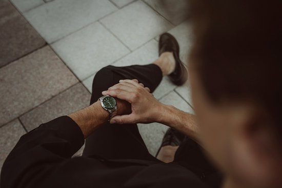 Een man draagt een zilverkleurig Edifice horloge met groene wijzerplaat, zittend met de handen op zijn schoot.