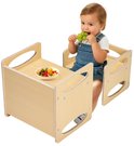 Een peuter met overall zit aan een naturel houten activiteitentafel met een stoel en eet druiven en een kleine salade.