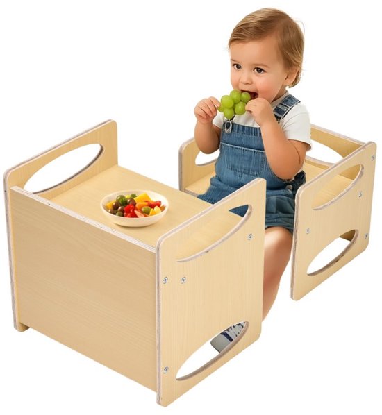 Een peuter met overall zit aan een naturel houten activiteitentafel met een stoel en eet druiven en een kleine salade.