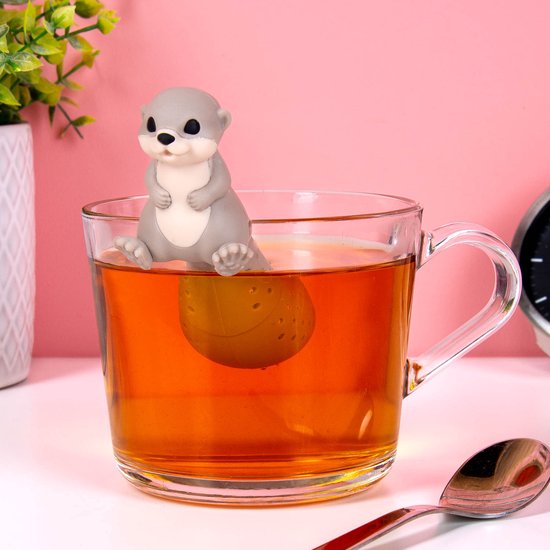 Grijze siliconen otter thee-ei in een glas warme thee. Een lepel ligt ernaast.
