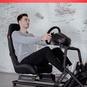 Man in grijze trui en zwarte jeans racet in een zwarte kuipstoel met racestuur en pedalen.