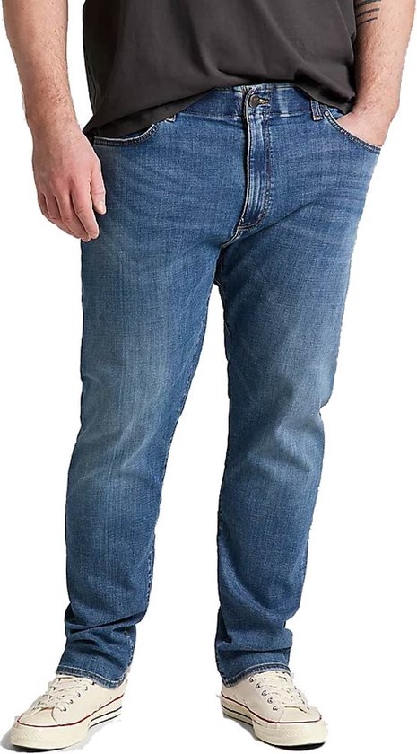 Man met een donkergrijs T-shirt en een lichtblauwe jeans, staand met handen in de zakken.