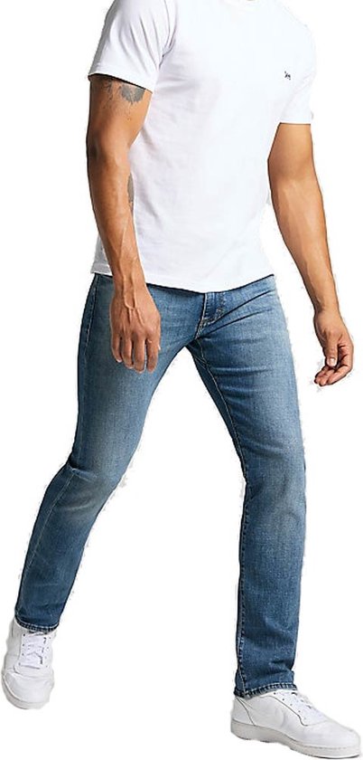 Man in een wit T-shirt en spijkerbroek loopt, met witte sneakers. De spijkerbroek is lichtblauw en slank.