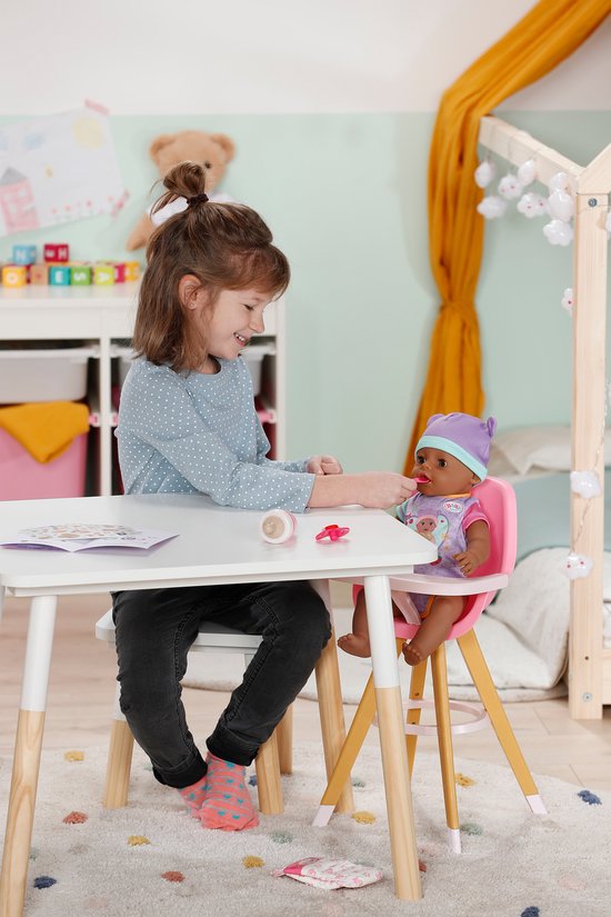 Meisje voert babypop aan tafel. De pop zit in een roze kinderstoel en draagt een paarse muts.