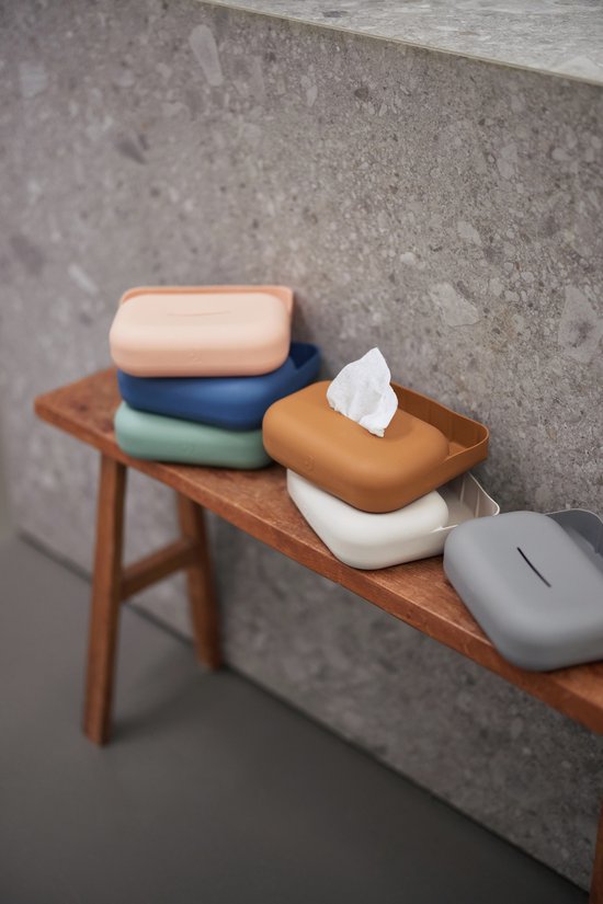 Stapel van verschillende kleuren siliconen doosjes voor billendoekjes op een houten bankje, één doosje met doekjes eruit.