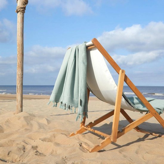 Jadegroene hamamdoek met witte strepen en kwastjes, hangend aan een houten strandstoel op een zonnig strand.