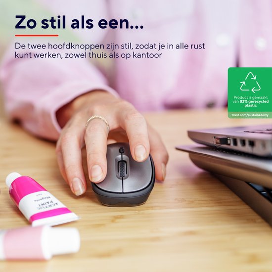 Hand die een stille, grijze draadloze Trust muis bedient, naast een laptop en verfbuisjes.