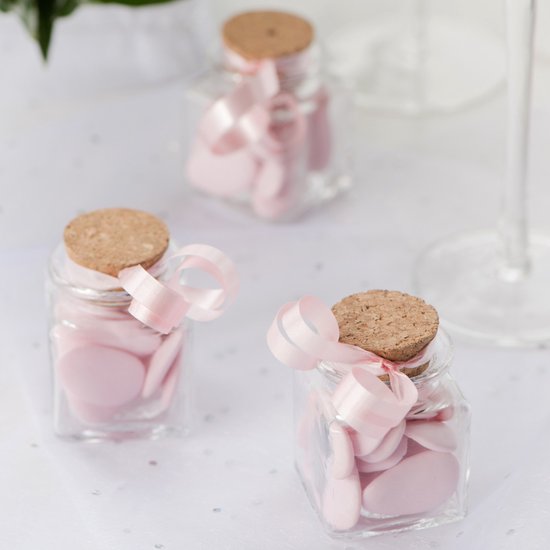 Trois petites bouteilles carrées en verre avec bouchon en liège, remplies de bonbons roses en forme de cœur, décorées de rubans roses.