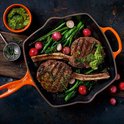 Oranje grillpan met twee grote karbonades, groenten, saus en kruiden, op een rustieke ondergrond.
