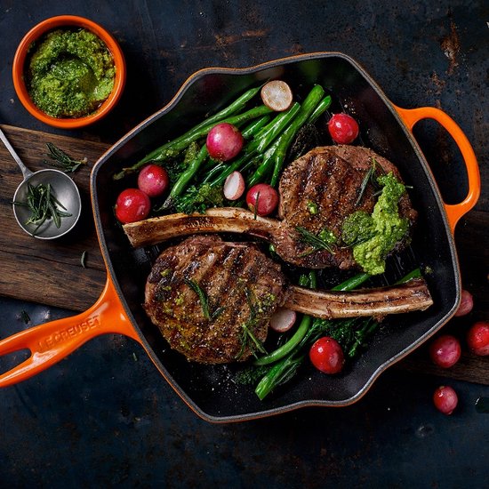 Oranje grillpan met twee grote karbonades, groenten, saus en kruiden, op een rustieke ondergrond.