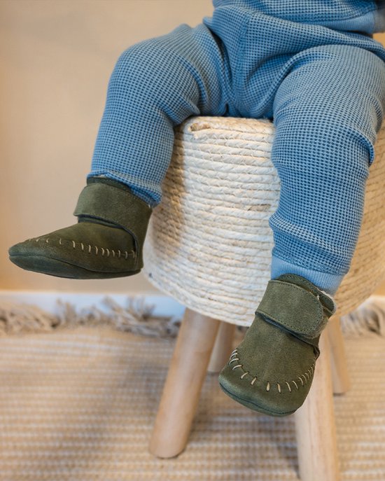 Groene suède babyslofjes met klittenband en gele stiksels, gedragen door een baby