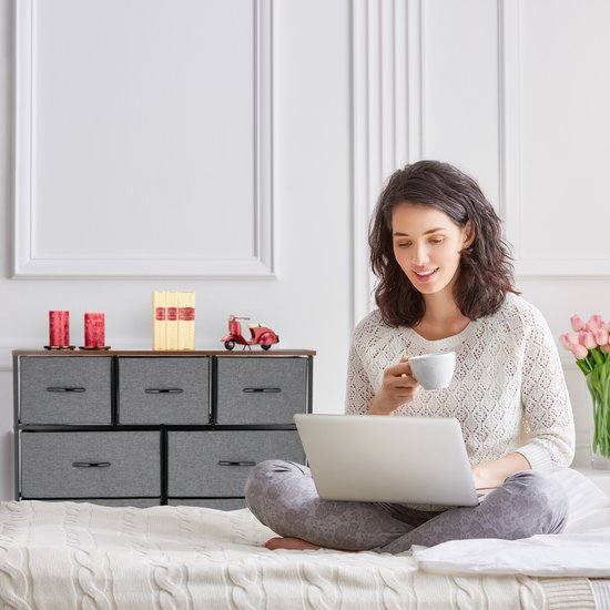 Dressoir met 6 grijze lades, houten top en zwart frame, naast een vrouw met laptop op bed.