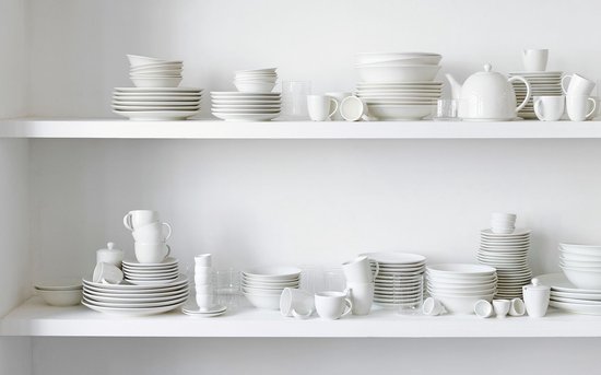 Een witte serviescollectie uitgestald op witte planken, met borden, kommen, mokken en theepotten.