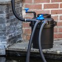 Zwarte vijverfilter met blauwe dop en handgreep, bij een stenen muur.