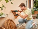 Man met bril schroeft een houten plank aan een muur met een groene Bosch boormachine.