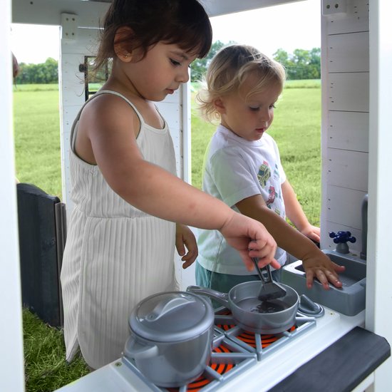 Twee kinderen spelen in de speelkeuken van het witte speelhuis. Meisje met jurk kookt, jongen met t-shirt kijkt toe.