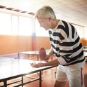 Grijsharige man met gestreepte trui en witte broek serveert een tafeltennisbal.