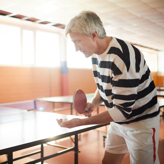 Grijsharige man met gestreepte trui en witte broek serveert een tafeltennisbal.