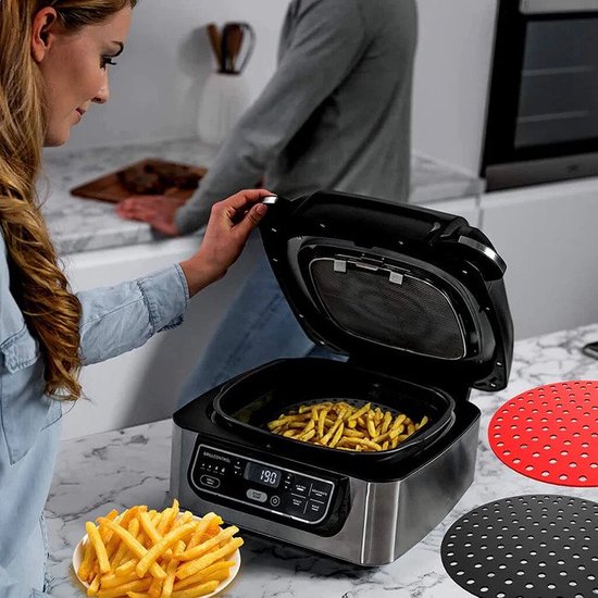 Een vrouw bereidt frietjes in een airfryer, met een rode en zwarte airfryer pad op het aanrecht.