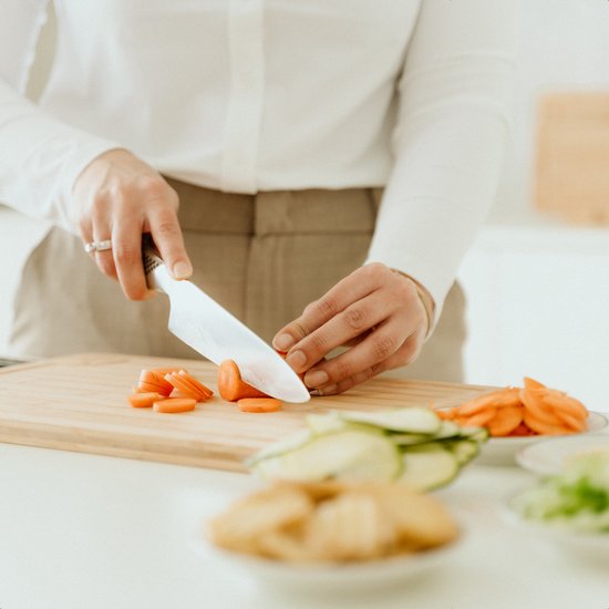 Gros plan sur une femme coupant des carottes sur une planche à découper en bambou. Des légumes et des herbes sont visibles.
