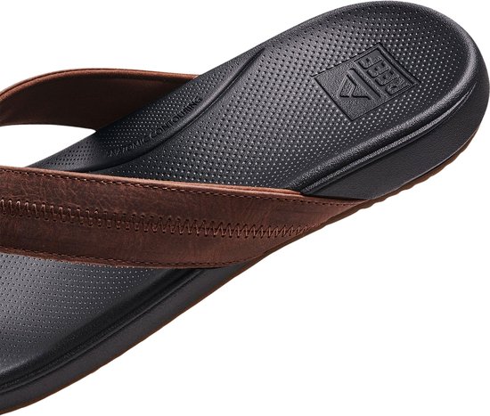 Detail van een bruine Reef slipper met zwart gestippeld voetbed, Reef logo en tekst 'ANATOMIC CONTOURING'.