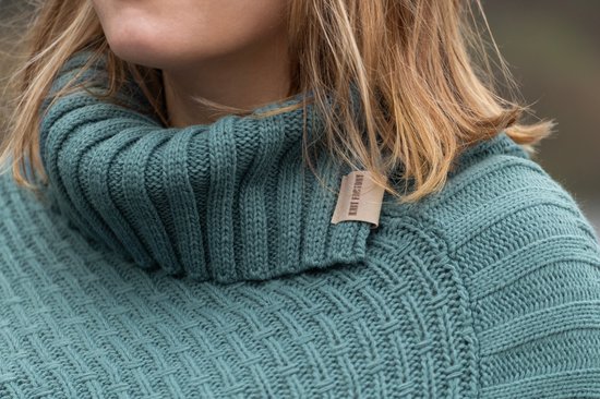 Close-up van een groenblauwe gebreide poncho met hoge ribbelkraag en een geborduurd label.