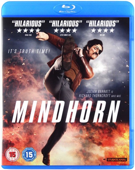 Man in bruin jack, grijze broek, met een ooglapje en snor, trapt in de lucht. Film: Mindhorn.
