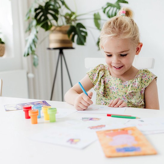 Een blond meisje zit aan een tafel en tekent met een blauwe stift, Peppa Pig stempels en potloden liggen op de tafel.